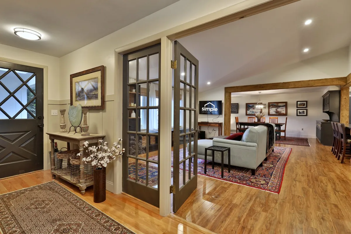 The view includes the entry area leading into a welcoming living room and adjacent dining area, all defined by inviting furniture, a plush rug, and large glass French doors that allow natural light to fill the space. The scene radiates comfort and sociability, highlighting settings for group gatherings and meals.