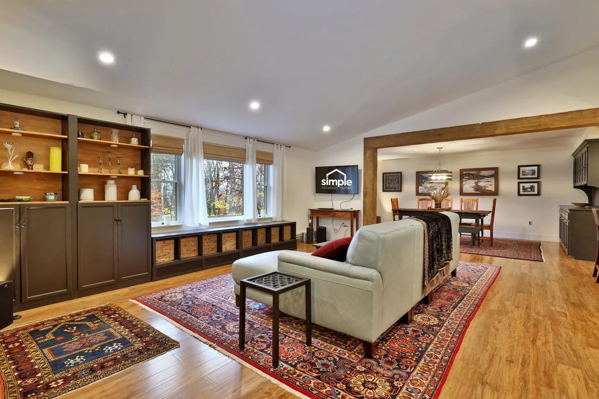 A sunlit living room featuring large windows with abundant natural light, a colorful area rug, a comfortable couch, and a bookshelf adorned with decorative items. The space feels open, relaxing, and conducive to both quiet moments and social gatherings.