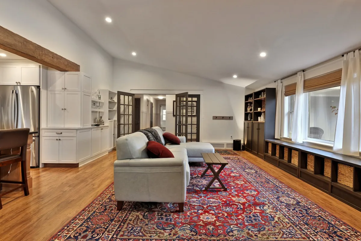 A comfortable and stylish living room featuring a plush sofa, a patterned rug, a coffee table, large windows allowing natural light to spill in, and a bookshelf. The setting feels inviting, perfect for relaxing, reading, or socializing in a bright atmosphere.