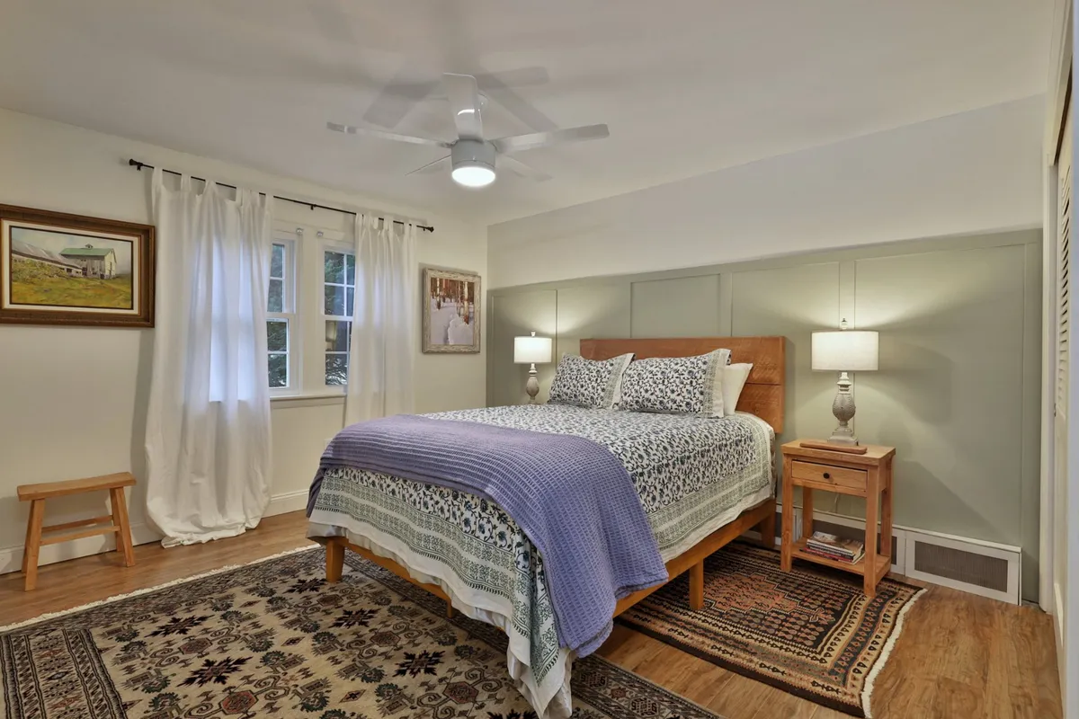 This image features a bedroom with patterned bedding, framed artwork on the wall, a wooden bedside table, and a window with curtains. Natural light streams in, creating a serene, restful atmosphere perfect for relaxing or enjoying a quiet morning coffee.