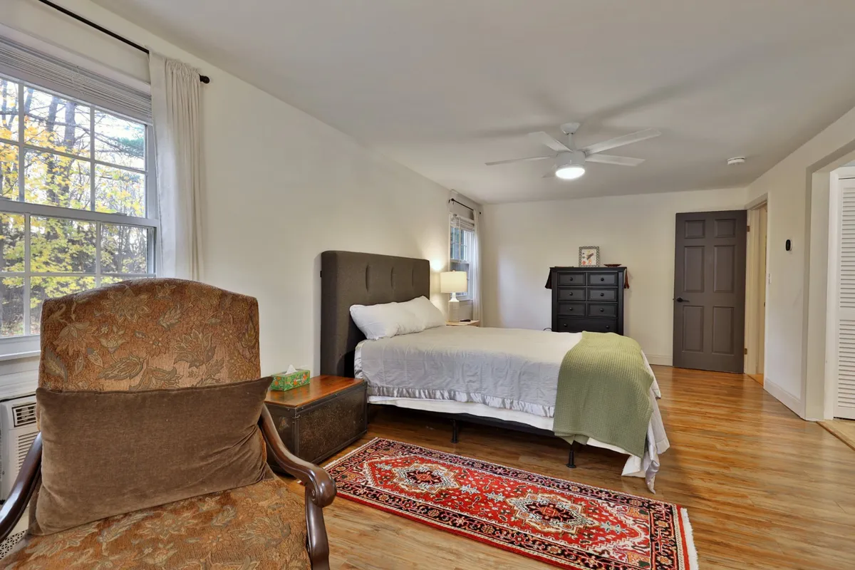 A tranquil bedroom scene showcasing a cozy bed, armchair, and dresser by a sunlit window, with soft lighting and a warm rug underfoot. The setting conveys peaceful mornings or restful evenings.