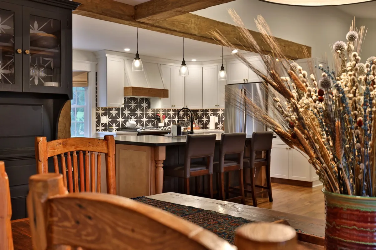 The image showcases a warm, open-concept dining and kitchen area featuring a large wooden dining table, a chic kitchen island, a decorative vase with dried flowers, and a striking geometric tile backsplash. The space is designed for gatherings, with plenty of natural light and a stylish, welcoming atmosphere perfect for sharing meals or entertaining.