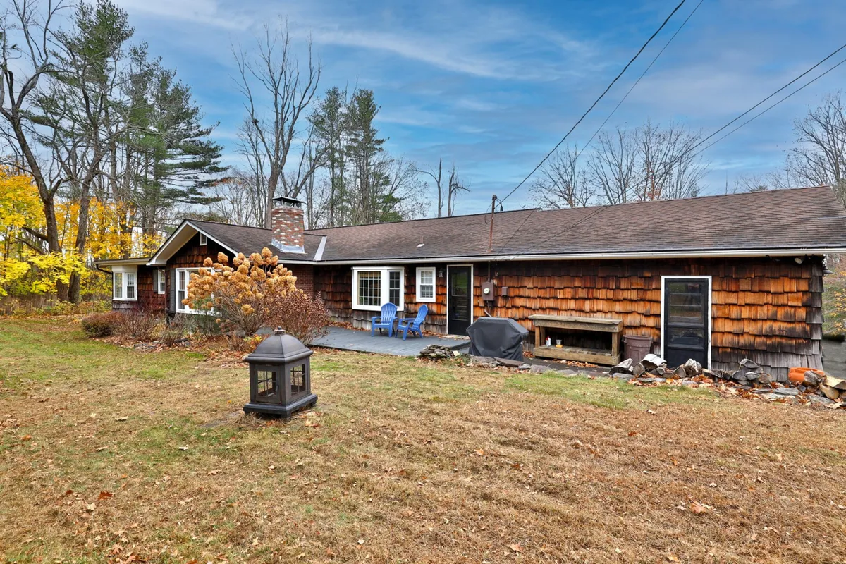 A rustic exterior with wooden shingles and inviting blue chairs positioned on a porch, set next to a garden area. The outdoor lantern suggests cozy evenings, while the visible fire pit area and garden seating make it ideal for relaxation and group gatherings.